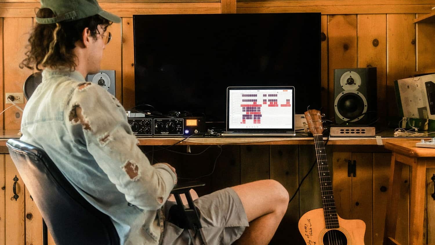 man sits dejectedly next to a desk with a computer on it. a guitar leans against the desk.