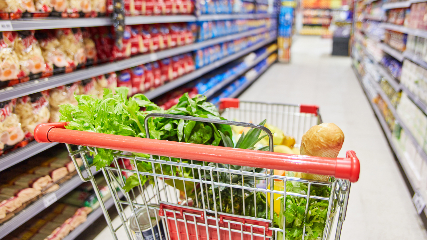 A shopping cart full of vegetables sits in the middle of a grocery store aisle.