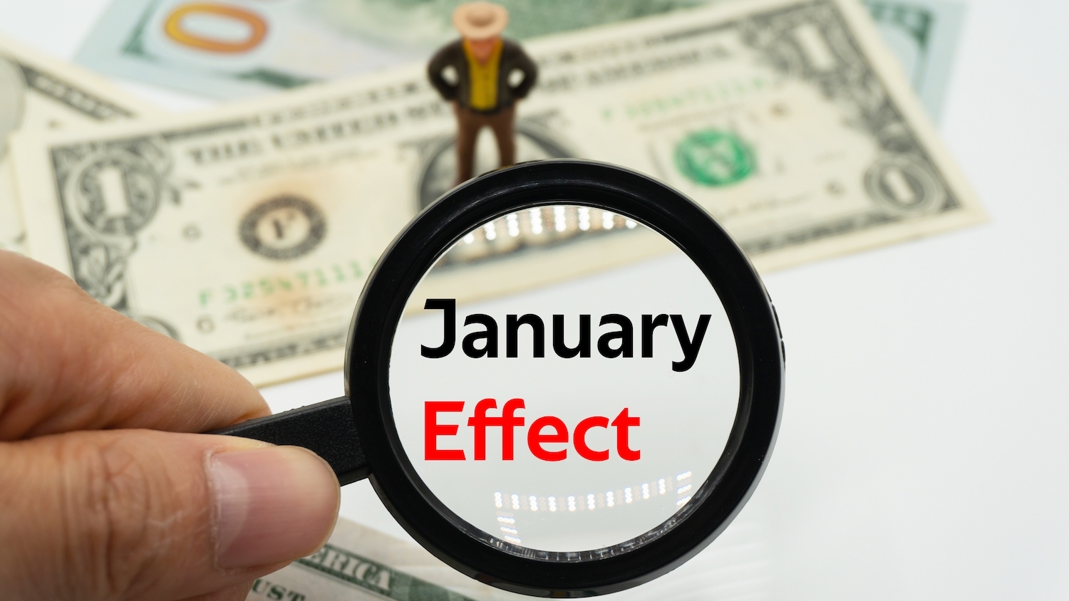 A magnifying glass held in two fingers enlarges the words "January Effect." One- and one-hundred-dollar bills are in the background, along with a miniature figure of a businessperson.